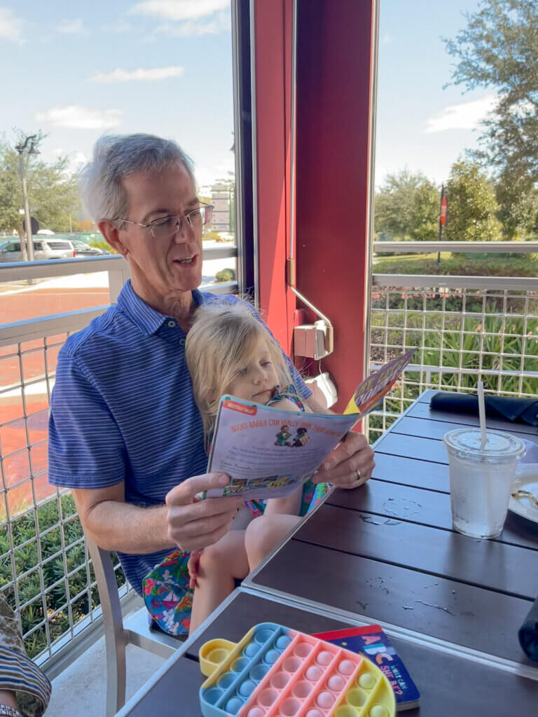 A grandfather reads a paperback book to a little girl at a restaurant table.  Other toys are in the foreground - a few mentioned as restaurant essentials.